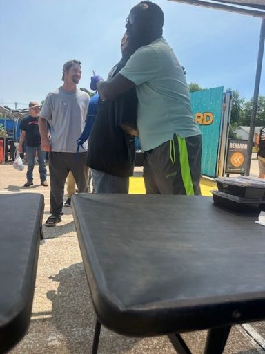 Two people embrace in a crowded outdoor setting while others stand nearby.